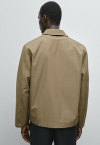 Lichte, bruine jas met een puntkraag, lange mouwen en drukkers aan de manchetten. Gladde textuur, minimalistisch ontwerp zonder zichtbare merknaam.