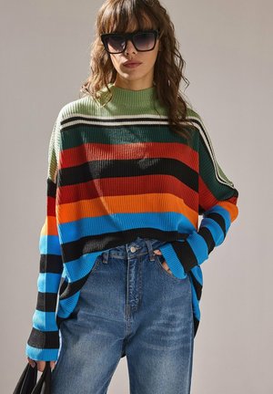 Femme portant un pull oversize rayé multicolore avec du vert, du rouge, de l’orange, du bleu et du noir, un jean bleu, de grandes lunettes de soleil noires et tenant un sac noir.