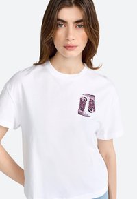 T-shirt en coton blanc avec un graphique floral rose et violet de bottes de cowboy sur la poitrine gauche. Manches courtes, col rond, coupe décontractée.