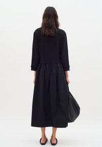 Schwarzes Kleid mit enganliegendem Oberteil und gerafftem Rock. Lange Ärmel, weiche Textur und eine lockere, fließende Form. Modell hält eine schwarze Tasche.