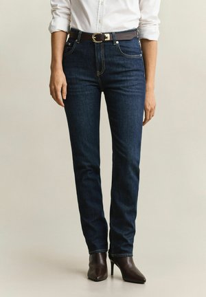 Person trägt dunkelblaue Jeans, braunen Ledergürtel, weißes langärmeliges Hemd mit aufgekrempelten Ärmeln und braune Stiefeletten mit Absatz, steht aufrecht.