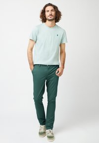 Lichtgroen katoenen T-shirt met een klein boompje-logo, gecombineerd met donkergroene chinos en grijs-groene sneakers, wat zorgt voor een casual look.