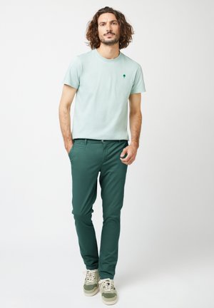Lichtgroen katoenen T-shirt met een klein boompje-logo, gecombineerd met donkergroene chinos en grijs-groene sneakers, wat zorgt voor een casual look.