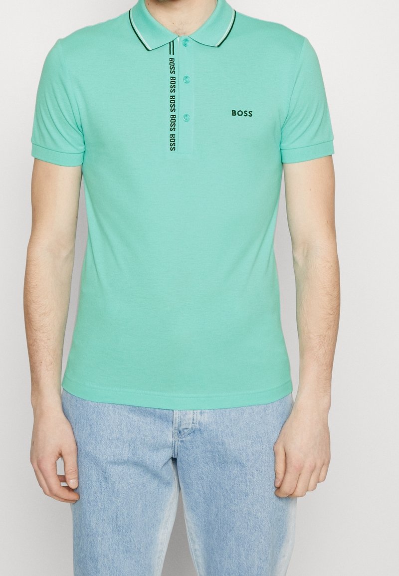 Homme portant un polo vert menthe avec le logo "BOSS" sur la poitrine et la patte de boutonnage, assorti à un jean bleu clair, sur un fond uni.