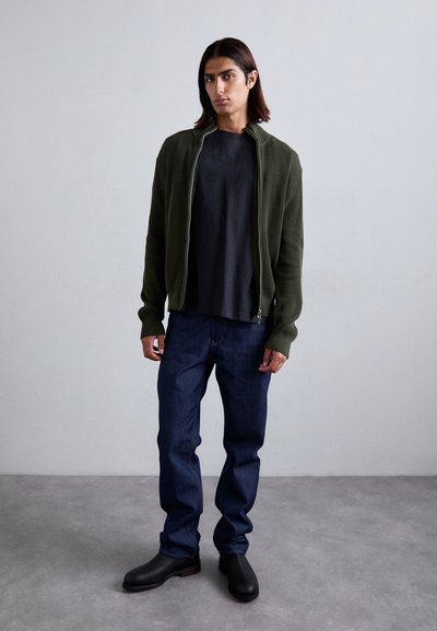 Jauns cilvēks ar vidēju garumu matiem, ģērbies tumši zaļā ar rāvējslēdzēju, melnā T-kreklā, tumši zilās biksēs un melnās zābakos, stāv iekštelpās.