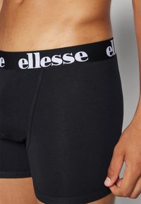 Schwarze Boxer-Shorts mit einem schwarzen elastischen Bund, der mit weißem "ellesse"-Logo versehen ist. Der Stoff wirkt glatt und enganliegend und reicht bis zur Mitte des Oberschenkels.
