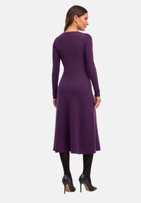 Abito a maglia viola a coste con maniche lunghe, lunghezza midi, vita aderente e gonna fluida. Abbinato a scarpe con tacco nere.