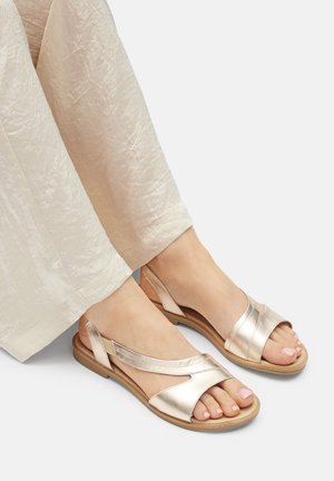 Metallisch goldene Sandalen mit offenem Zehenbereich und schlankem Design, ausgestattet mit gekreuzten Riemen und einer niedrigen, flexiblen Sohle. Kombiniert mit hellfarbenen, strukturierten Hosen.