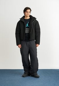 Giacca a vento nera con chiusura a zip, indossata sopra una maglietta nera con strisce bianche e verde acqua. Pantaloni neri larghi e calzature nere.