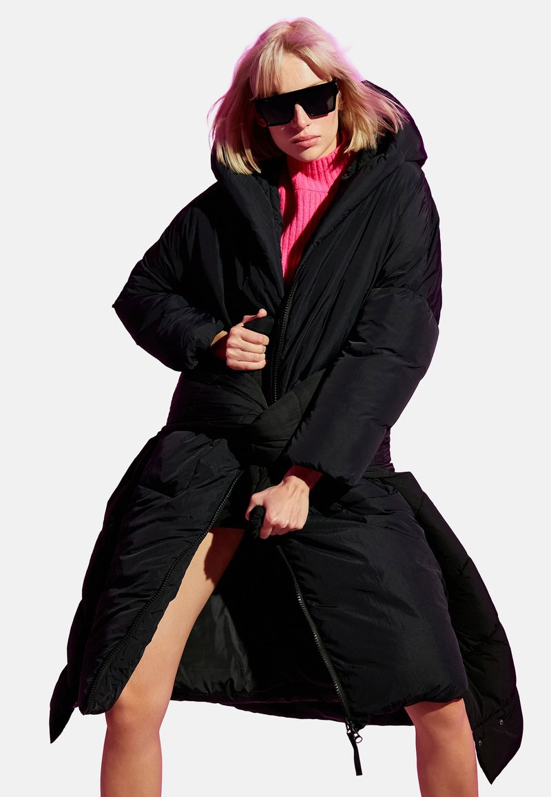 Giacca a vento imbottita realizzata in tessuto nero, con un ampio cappuccio, silhouette oversize e zip frontale con lacci in vita per la chiusura.