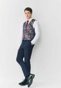 Navyfarbener Weste mit Blumenmuster, ergänzt durch burgunderfarbene Akzente, kombiniert mit einem weißen Hemd und dunklen Hosen. Schwarze Lederschuhe runden den Look ab.