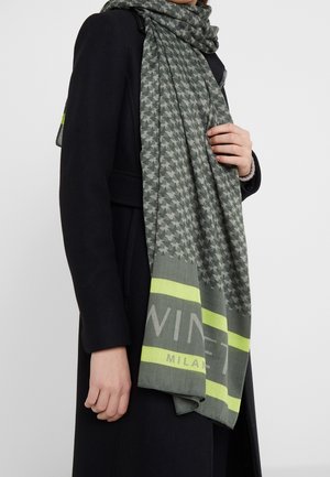 Personne portant un manteau noir et une écharpe verte à motifs avec des rayures jaunes et le texte "Milano", tenant l'écharpe près de la poitrine.