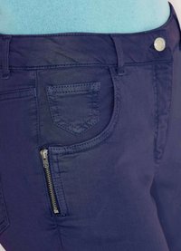 Close-up van marineblauwe broek met een voorzak, kleine ritsdetail, knop en riemlussen, gedragen met een lichtblauwe top.