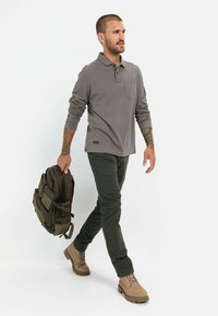 Grijze lange mouw polo met een borstzak, groene broek en beige laarzen. Een donkere rugzak wordt in de hand gehouden.