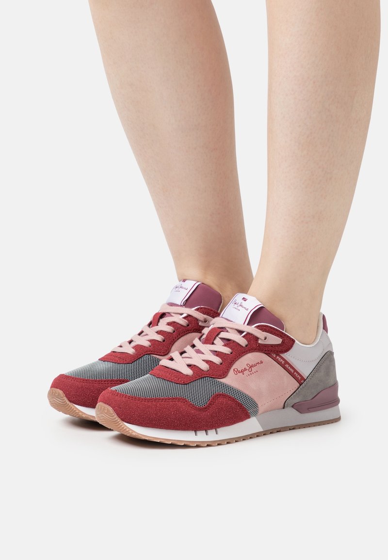 Pieds portant des baskets Pepe Jeans rouges, roses, grises et blanches avec des lacets sur un fond blanc.