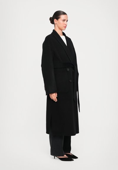 Musta villa wrap-takki, jossa on shaalikaulus, napit edessä ja sivutaskut. V Wear valkoisen paidan päällä, yhdistetty tummiin farkkuihin ja mustiin korkokenkiin.