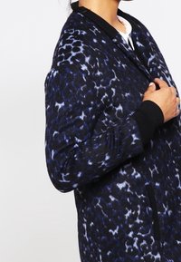 Donna che indossa un cappotto leopardato blu scuro e nero con polsini neri, mentre tiene il colletto del cappotto con una mano.
