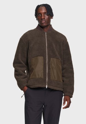 Homme portant une veste polaire marron foncé zippée avec de grandes poches avant et un pantalon noir, debout devant un fond uni.