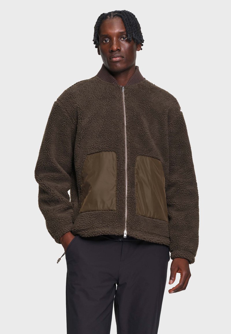 Homme portant une veste polaire marron foncé zippée avec de grandes poches avant et un pantalon noir, debout devant un fond uni.
