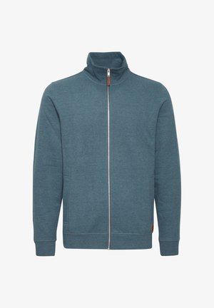 Tsinzipperiga dressipluus türkiissinises värvitoonis, valmistatud pehmest kangast. Omab kõrget kraed, pikki varrukaid ja kahte küljetaskut. Minimalistlik disain, ilma mustriteta.