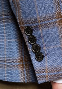 Niebiesko-brązowa tkanina w kratę o teksturowym wykończeniu; zawiera cztery czarne guziki na mankiecie. Dziurki na guziki są starannie obszyte.
