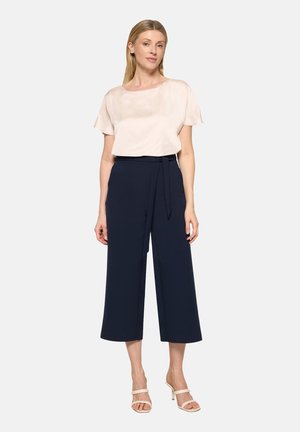 Woman wearing short-sleeve beige blouse, high-waisted navy wide-leg cropped pants with belt, and beige heeled sandals standing against white background.