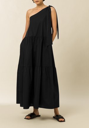 Woman wearing a long black one-shoulder dress with a tied strap and black slide sandals standing against a plain background.