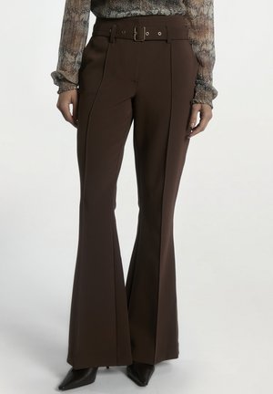 Vrouw die hooggesneden donkerbruine uitlopende broek draagt met riem en puntige zwarte schoenen, gecombineerd met een getextureerde top met lange mouwen.