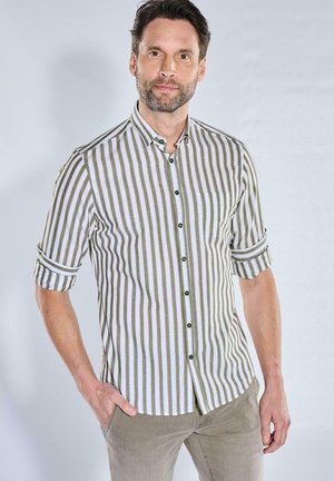 Mann mit kurzen dunklen Haaren und Bart, der ein grün-weiß vertikal gestreiftes Hemd mit hochgekrempelten Ärmeln und beigen Hosen trägt, steht mit einer Hand in der Tasche.