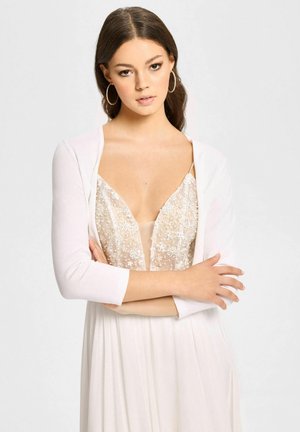 Ung kvinde med mørkt hår iført store hoop-øreringe, en gennemsigtig paliet-top og en hvid cardigan, med armene krydset foran en ensfarvet baggrund.