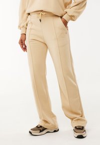 Béžové tepláky z měkké, texturované látky s elastickým pasem na šňůrku, boční kapsy a svislé švová detaily pro zvýraznění stylu.