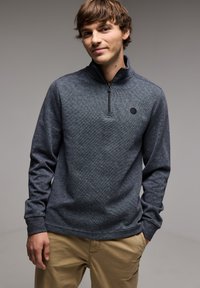 Pullover grigio scuro con colletto a zip a quarti, tessuto texture e un piccolo logo rotondo sul petto. Maniche lunghe e vestibilità morbida.