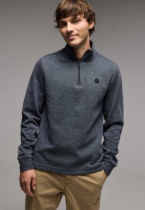 Donkergrijze pullover met een kwart-rits kraag, gestructureerde stof, en een klein rond logo op de borst. Lange mouwen en een ontspannen pasvorm.