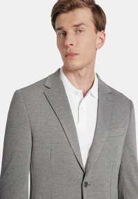 Szary żakiet w wzór houndstooth z klapami, na jeden guzik i kieszenią na piersi, nałożony na białą koszulkę polo. Gładka faktura materiału.