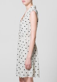 Abito bianco senza maniche con motivi a cuore neri, tessuto morbido, vita aderente e colletto, tagliato sopra il ginocchio per un look sartoriale.