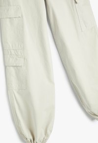 Cargo-byxor i ljusbeige tyg med flera sidofickor, elastiska mudd, och en avslappnad passform med synliga sömnadsdetaljer.