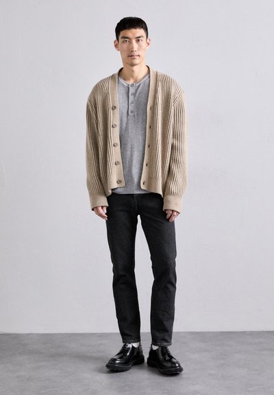 Beige ribbet cardigan med store knapper over en lys grå henley-skjorte, sammen med svarte jeans og svarte snøresko.