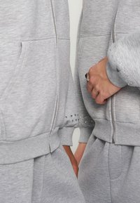 Gros plan de deux personnes portant des sweats à capuche zippés gris clair et des pantalons de survêtement assortis, une main visible avec une bague en argent, manches des sweats avec de petites décorations.
