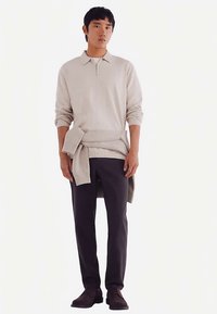 Ljusbeige tröja med zip-up-krage, bärs över en matchande stickad omslagskofta. Mörka byxor och bruna skor kompletterar outfiten. Minimalistisk design.