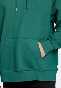 Grön huvtröja med en framficka, ribbade mudd och nederkant, samt en diskret broderad logga ovanför fickan. Slät tygtextur.