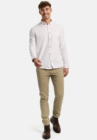 Weißes Hemd mit Knöpfen, beige Chinos, braune Schnürstiefel. Das Hemd hat einen Kragen und knöpfe in Holzoptik, mit hochgekrempelten Ärmeln für einen lässigen Look.