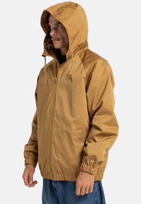 Bruine zip-up jack met een capuchon, elastische manchetten en een voorzak. Gemaakt van gladde stof, met een subtiel logo op de borst.