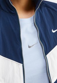 Námořnická a bílá větrovka se stříbrným zipem, na které je logo Nike. Pod ní je světle modré tričko, také zdobené malým logem Nike.