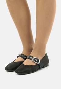 Bailarinas de ante negro con punta afilada y una correa ancha con herrajes de plata. La suela es plana y ofrece un diseño minimalista.
