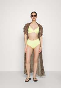 Biquíni amarelo com bordas em forma de ondas, combinado com um kaftan de padrão leopardo castanho. A modelo usa óculos de sol pretos e chinelos.