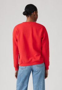 Red sweatshirt with a crew neck and ribbed cuffs, made of soft fabric. Paired with light blue jeans, showcasing a relaxed fit.