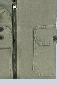 Green zippered jacket detail featuring a textured fabric, a chest pocket with a flap and button, and a smooth zipper outlined in green.
