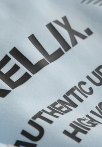 Lichtblauwe stof met grote, vetgedrukte zwarte tekst. De textuur lijkt glad en licht reflecterend, met een nadruk op grafische ontwerpelementen.