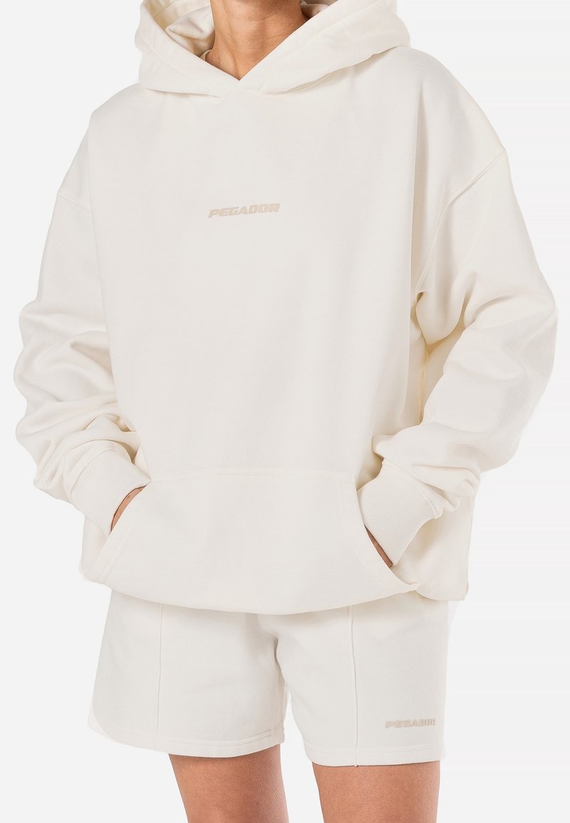 Sweat à capuche couleur crème en tissu doux avec une poche avant et des poignets côtelés, assorti à un short assorti avec un branding discret.