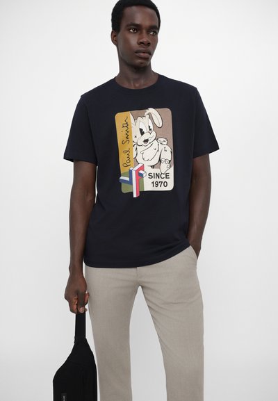 T-shirt navy avec un imprimé de lapin en cartoon dans un design rectangulaire, comportant le texte "Paul Smith" et "Depuis 1970". Le modèle tient un sac noir.
